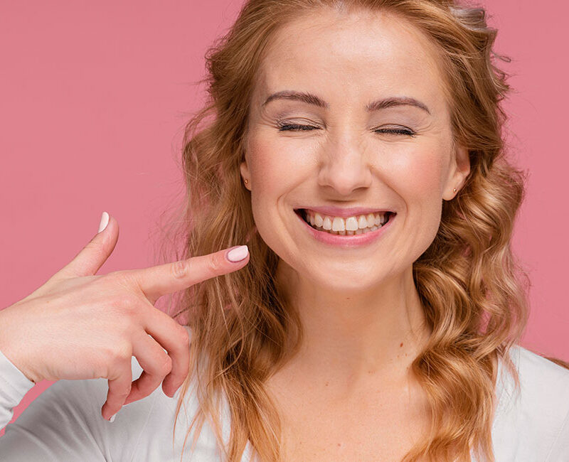 A woman is pointing to her teeth on a pink background, demonstrating how clear aligners work.