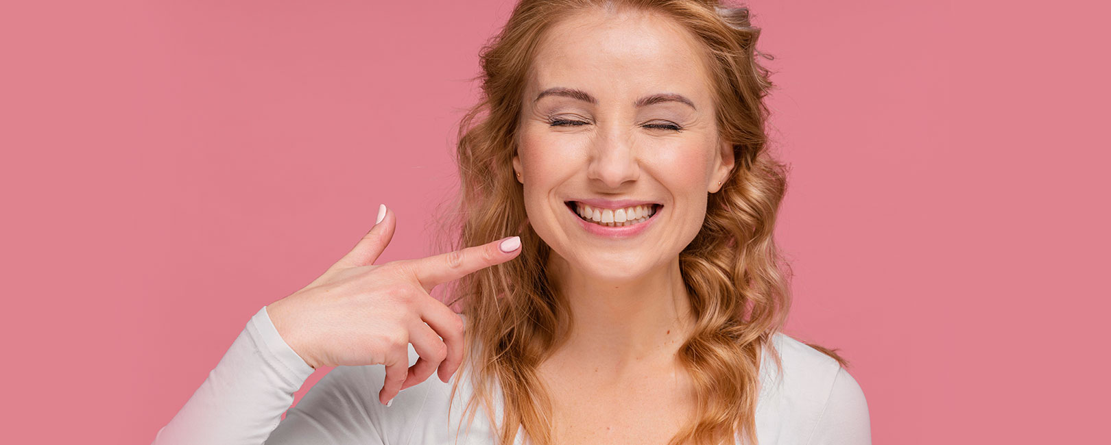 A woman is pointing to her teeth on a pink background, demonstrating how clear aligners work.