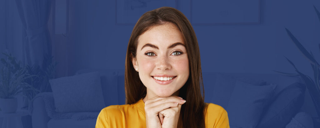 A woman smiling in front of a blue living room, confidently showcasing her straightened smile achieved from the comfort of her own home using teeth straightening kits.